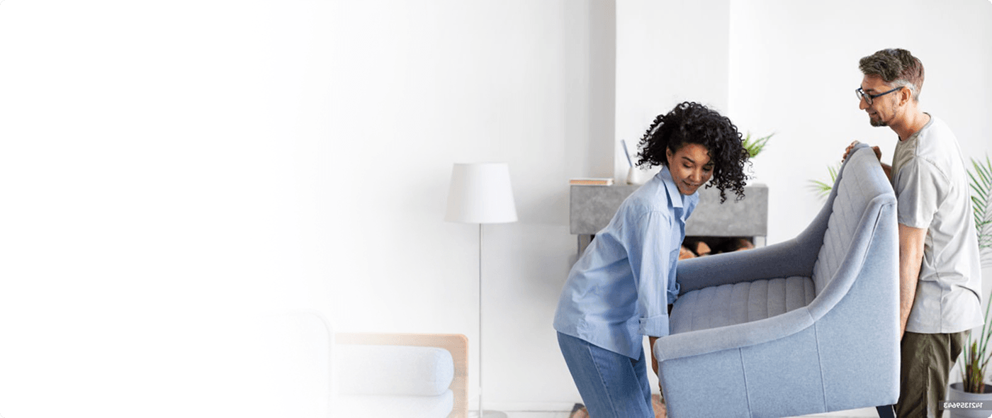 A man and woman carry a large chair through the living room of a home.
