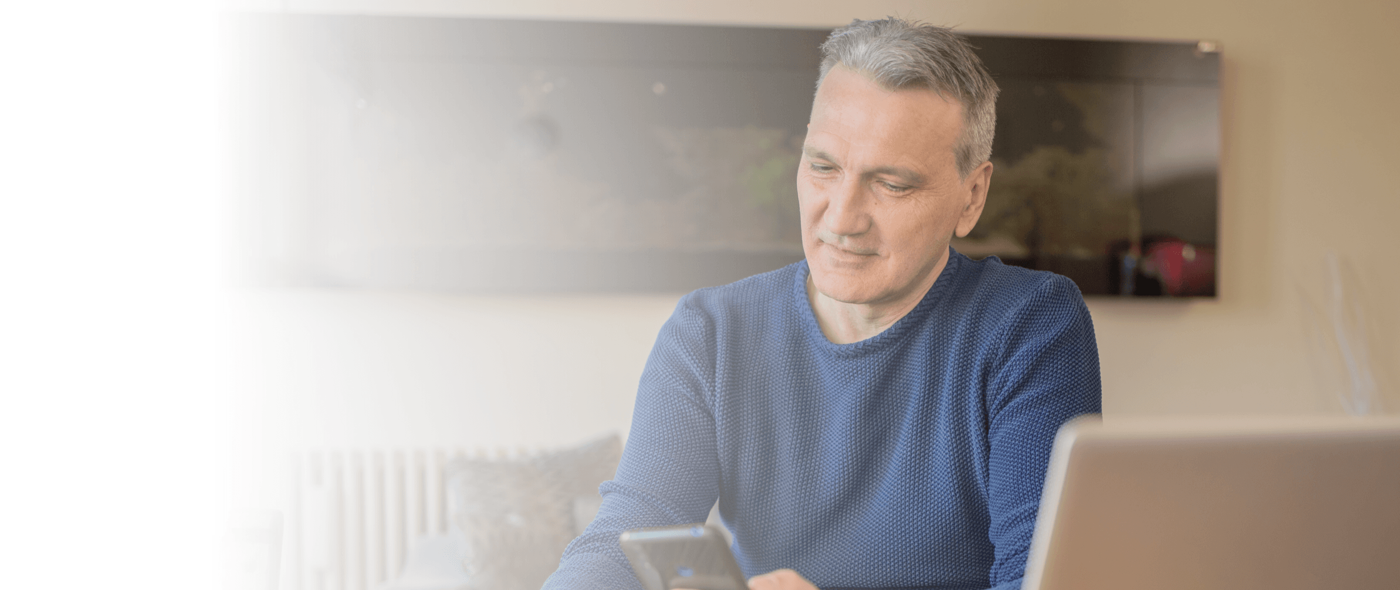 A man sits at a table with a computer holding a smartphone.